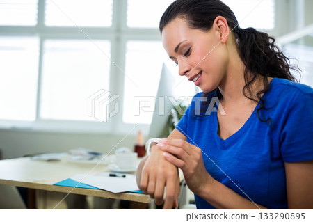 Woman sitting at desk adjusting smartwatch by window with monitor, plant, copy space Woman sitting at desk adjusting smartwatch by window with monitor, plant, copy space 133290895