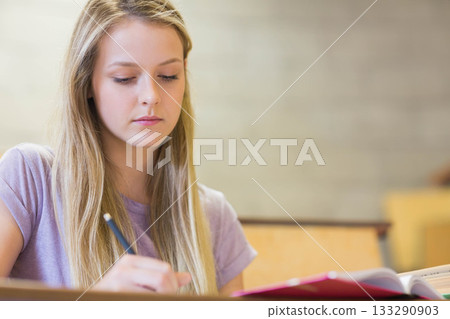 Teenage girl in lilac t-shirt writing notes in notebook at classroom desk with pencil, copy space 133290903