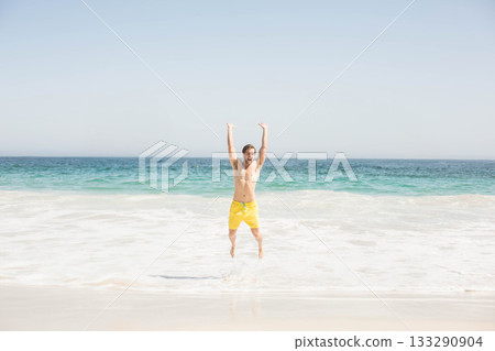 Man jumping in shallow surf on sandy shore wearing yellow swim trunks with ocean waves 133290904