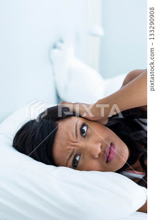 African American woman lying on white pillow on bed in bedroom pressing hand against ear African American woman lying on white pillow on bed in bedroom pressing hand against ear 133290908