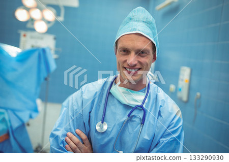 Mid adult male surgeon wearing scrubs crossing arms by OR table under OR lights with stethoscope Mid adult male surgeon wearing scrubs crossing arms by OR table under OR lights with stethoscope 133290930