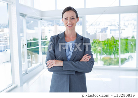 Businesswoman standing with arms crossed and smiling in office with glass walls and city skyline 133290939