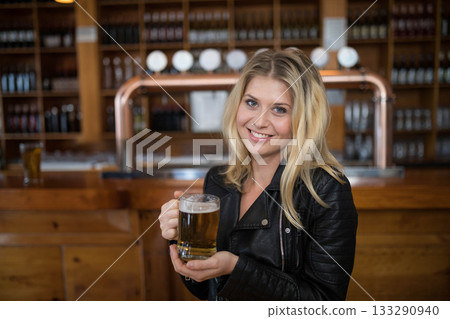 Woman in her twenties holding beer mug smiling toward camera at metal beer taps in bar Woman in her twenties holding beer mug smiling toward camera at metal beer taps in bar 133290940