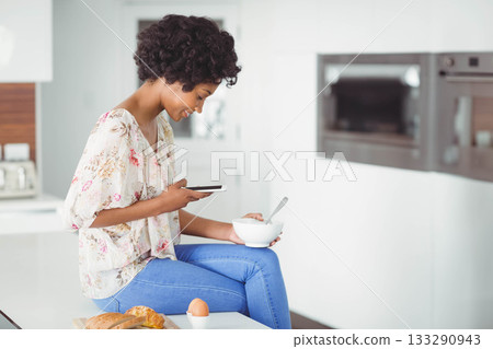 African American woman sitting on kitchen countertop holding smartphone and bowl in kitchen at home African American woman sitting on kitchen countertop holding smartphone and bowl in kitchen at home 133290943