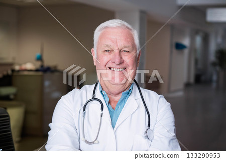 Senior male doctor standing in hospital reception smiling while wearing lab coat with stethoscope Senior male doctor standing in hospital reception smiling while wearing lab coat with stethoscope 133290953