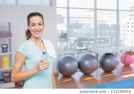 Female fitness trainer standing near windows holding water bottle and towel in studio, copy space 133290958