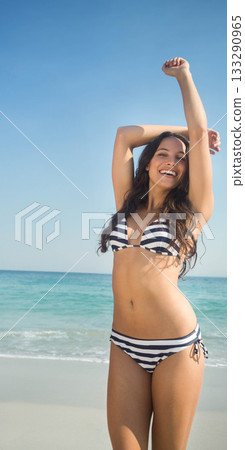 Woman standing on sandy beach raising arms under clear blue sky wearing striped bikini, copy space Woman standing on sandy beach raising arms under clear blue sky wearing striped bikini, copy space 133290965