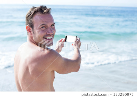 Mid-adult man smiling while holding smartphone with blank screen at water's edge on sandy beach Mid-adult man smiling while holding smartphone with blank screen at water's edge on sandy beach 133290975
