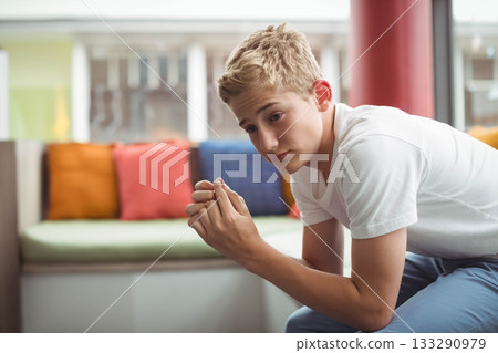 Teen boy leaning and gazing on cushioned bench by large window with colorful pillows, red column 133290979
