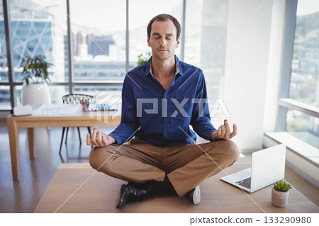 Mid-adult man meditating cross-legged on wooden desk in open-plan office with laptop and plants 133290980