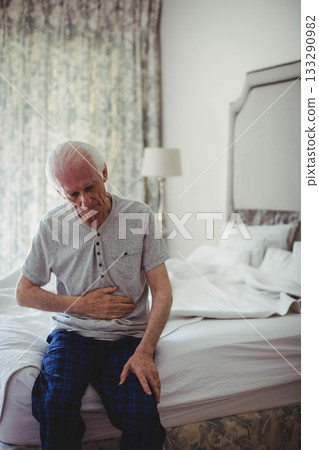 Senior man sitting on edge of unmade bed in softly lit bedroom with floral curtain, lamp Senior man sitting on edge of unmade bed in softly lit bedroom with floral curtain, lamp 133290982