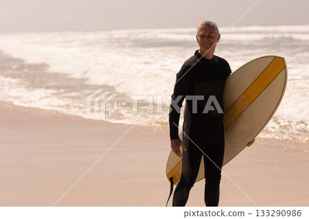 Senior man standing knee-deep at beach holding surfboard and wearing wetsuit, copy space 133290986