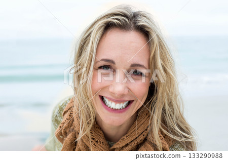 Blonde woman smiling at camera while standing on sandy beach wearing brown knit scarf green sweater Blonde woman smiling at camera while standing on sandy beach wearing brown knit scarf green sweater 133290988