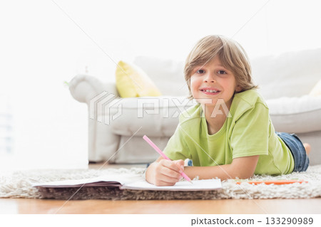 Boy lying in living room writing in notebook with pink pencil and coloring sheets, copy space 133290989