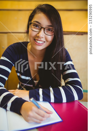 Asian female student writing notes with pen in notebook at lecture hall desk with pink binder Asian female student writing notes with pen in notebook at lecture hall desk with pink binder 133290995