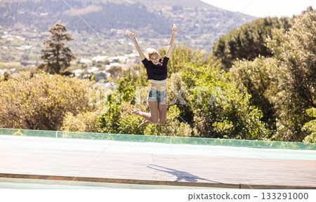 Woman leaping above swimming pool on wooden deck past glass railing overlooking hillside houses 133291000