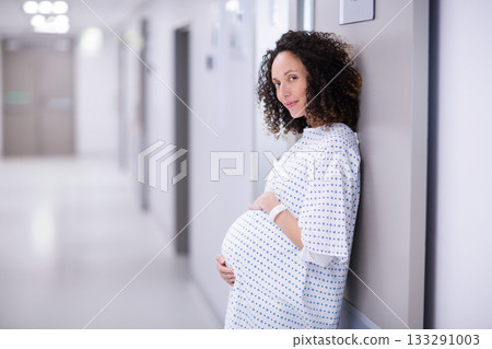 Pregnant woman leaning in hospital corridor wearing gown and wristband, cradling belly, copy space 133291003