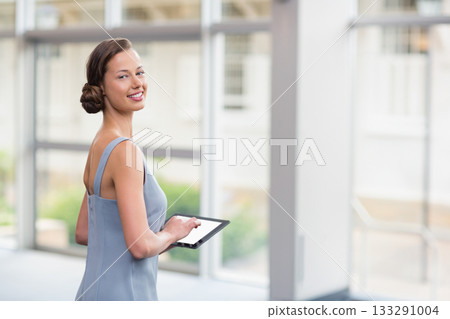 Female professional in blue dress holding tablet while looking back in office corridor, copy space 133291004