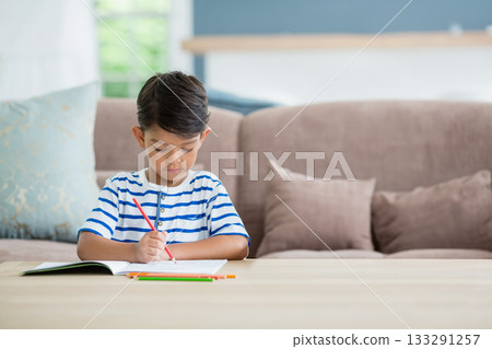 Asian boy holding red pencil while coloring notebook at wooden coffee table in living room 133291257