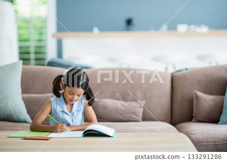 Child writing in open notebook with colored pencils at wood coffee table in living room Child writing in open notebook with colored pencils at wood coffee table in living room 133291286
