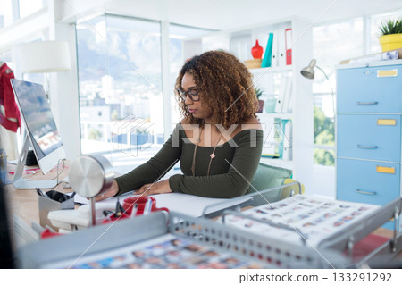 African American woman working at desk in studio reviewing photographic prints on computer monitor 133291292