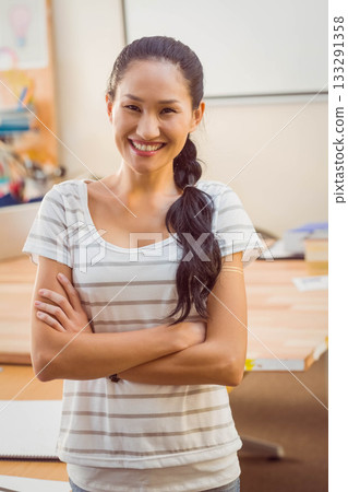 Asian woman standing crossing arms and smiling at classroom desk with whiteboard and art supplies 133291358