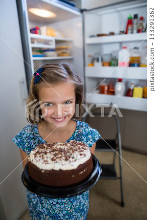 Smiling girl in blue dress holding white-frosted chocolate cake near open refrigerator in kitchen 133291362