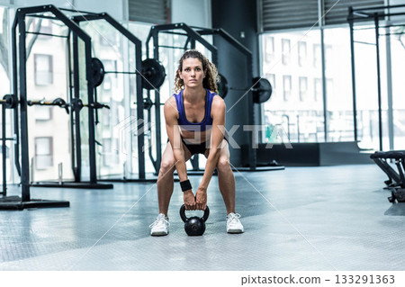 Woman performing kettlebell squat in gym area in sportswear with squat racks and weight plates Woman performing kettlebell squat in gym area in sportswear with squat racks and weight plates 133291363
