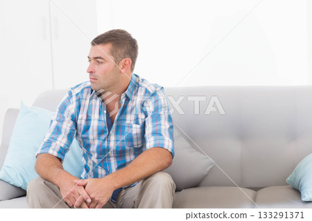 Man sitting on light grey sofa at home gazing left with pillows and cabinets, copy space 133291371