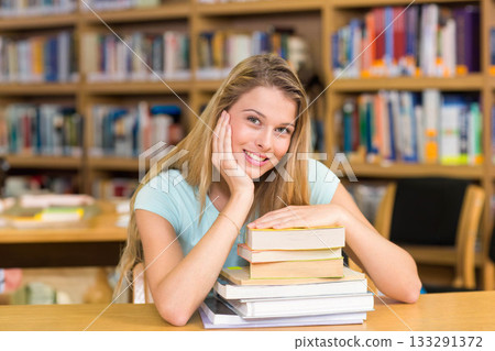 Teen girl leaning forward at wooden table in library with bookshelves and chairs reading textbooks 133291372