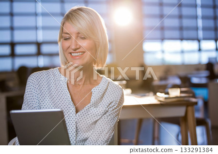 Smiling woman holding tablet, sitting at desk in office with grid windows, light blouse, coffee cup 133291384