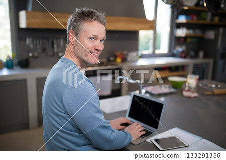 Mature male sitting at kitchen island, working on laptop with tablet, documents and mixing bowl Mature male sitting at kitchen island, working on laptop with tablet, documents and mixing bowl 133291386