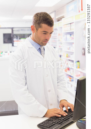 Mid adult male pharmacist typing on keyboard behind counter at pharmacy with medication shelves 133291387