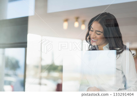 Woman standing behind glass partition leaning forward and smiling in office with lights, copy space Woman standing behind glass partition leaning forward and smiling in office with lights, copy space 133291404