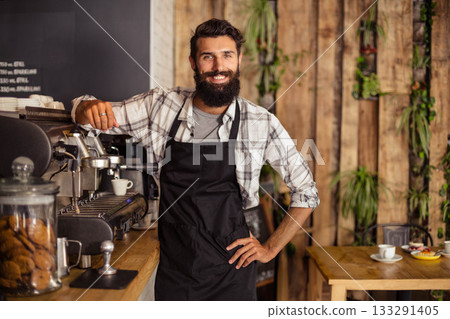 Male barista in black apron leaning on wooden counter at cafe preparing espresso with cookie jar Male barista in black apron leaning on wooden counter at cafe preparing espresso with cookie jar 133291405