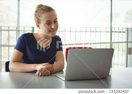 Teenage girl typing on silver laptop at study area by metal window railings with red chair Teenage girl typing on silver laptop at study area by metal window railings with red chair 133291407