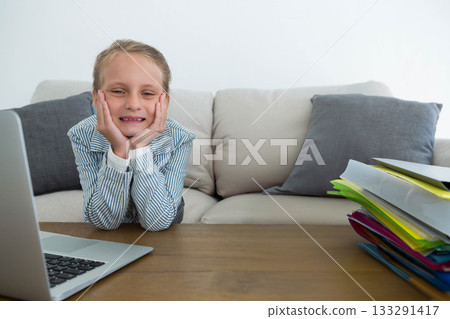 Girl resting chin on hands smiling at wooden table with silver laptop and colorful folders 133291417
