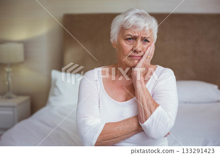 Senior woman sitting on bed in bedroom pressing hand against cheek beside nightstand lamp 133291423