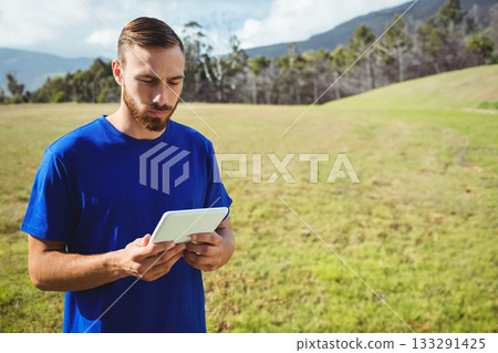 Man standing in grassy meadow holding white tablet and looking down at screen, copy space 133291425
