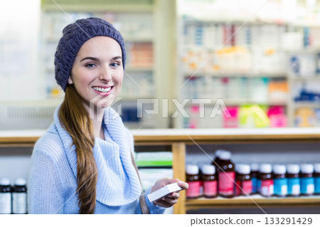 Teenage girl standing at pharmacy counter holding medicine box and smiling in beanie and sweater 133291429