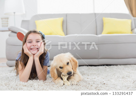 Girl child lying on living room rug smiling while puppy chewing rope toy, copy space 133291437