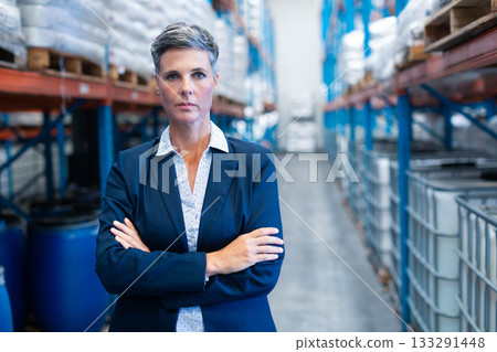 Female warehouse manager wearing blazer standing with arms crossed in aisle among pallets and drums 133291448