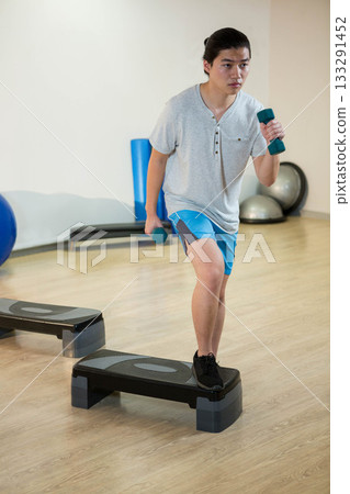 Asian woman stepping onto platform in gym holding teal dumbbells wearing grey shirt, blue shorts 133291452