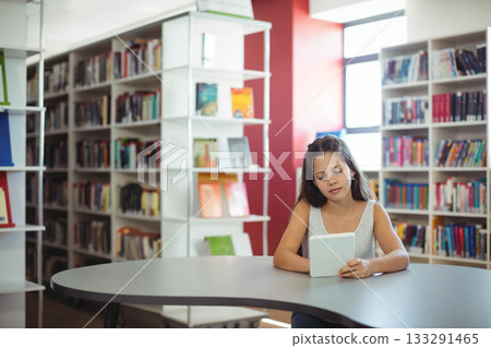 Teenage girl sitting at curved gray library table reading tablet by bookshelves, window, copy space 133291465
