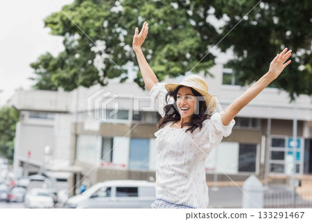 Female tourist raising arms overhead on sidewalk in front of modern building wearing straw sun hat Female tourist raising arms overhead on sidewalk in front of modern building wearing straw sun hat 133291467