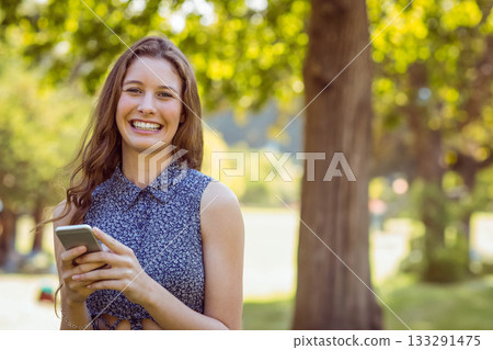 Teenage girl standing in sunlit leafy park clearing wearing floral crop top and holding smartphone Teenage girl standing in sunlit leafy park clearing wearing floral crop top and holding smartphone 133291475