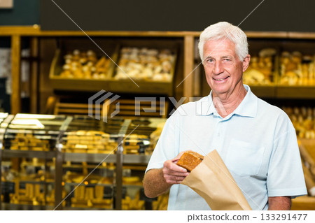 Senior man smiling and holding loaf of bread in paper bag inside bakery shop, copy space 133291477