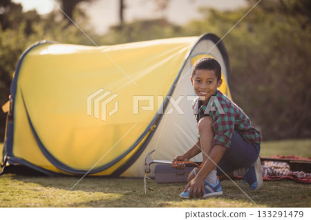 African American boy kneeling at park camp using hammer securing stake by yellow tent, copy space African American boy kneeling at park camp using hammer securing stake by yellow tent, copy space 133291479