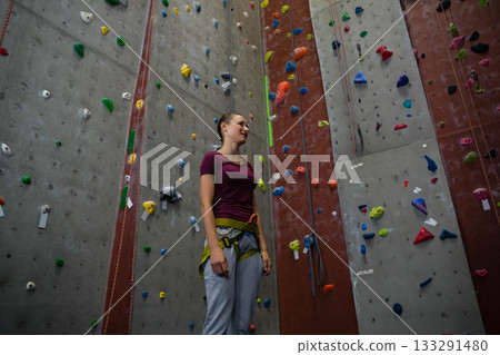 Female climber standing at rock climbing gym wearing maroon top and harness gripping colorful holds Female climber standing at rock climbing gym wearing maroon top and harness gripping colorful holds 133291480