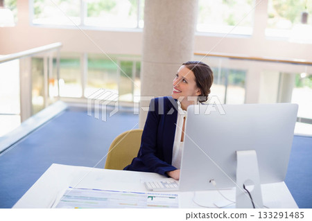 Female office worker wearing navy blazer smiling, looking up at monitor at office desk, copy space 133291485